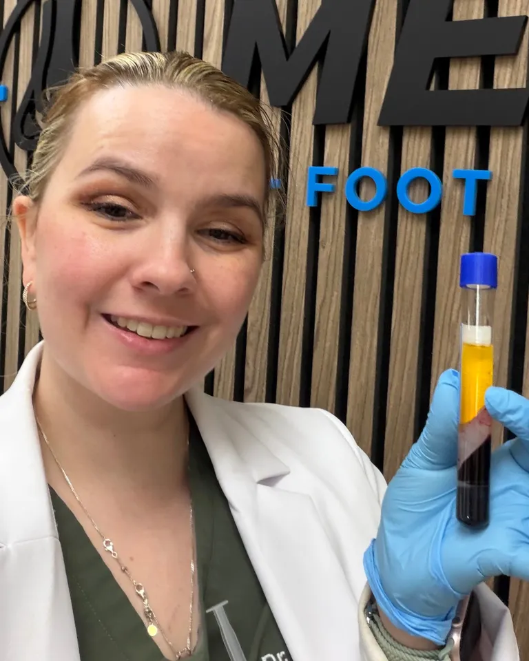 Dr. Kayla Wright, wearing a white coat and blue exam gloves, holds up a small tube of blood separated into layers, with the pale platelet-rich plasma on top, in front of the Metro Foot & Ankle logo wall.